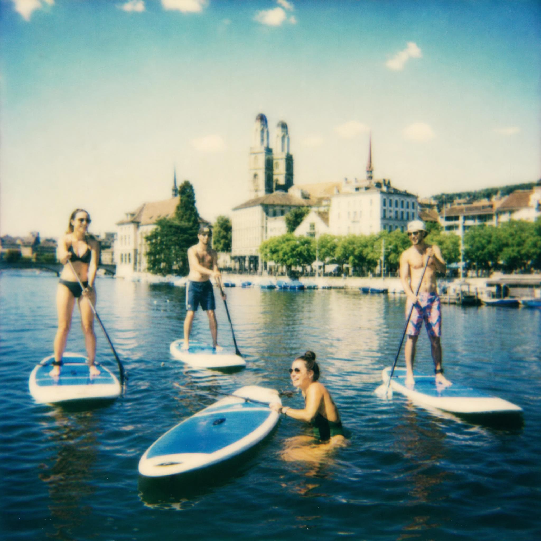 Eine Stand-up-Paddleboard-Gruppe auf dem Zürichsee – eine Person kühlt sich im Wasser neben ihrem Board ab, im Hintergrund die Zwillingstürme des Grossmünsters